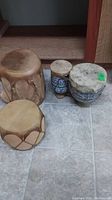 Four hand drums on tiled floor: two larger tan wooden drums with rope ties and two smaller ceramic base drums with painted designs.