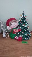 Front view of elf figurine hugging decorated Christmas tree with attached electrical cord, showing colorful bulbs and white snow accents.