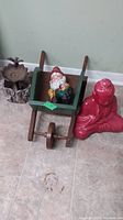 Ceramic elf figurine sitting inside a green wooden wheelbarrow with brown handles and wheel, wooden bird feeder on left side, and red Buddha statue on the floor to the right.