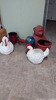 Shows all items in the lot including the two white chicken planters, teddy bear planter, metal tractor planter, and blue gazing ball on tiled floor.