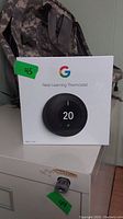 Front view of sealed white box featuring Google Nest Learning Thermostat with temperature set to 20 degrees shown on device face.