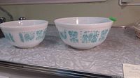 Two Pyrex Amish Butterprint mixing bowls side by side, showing similar teal designs on white milk glass.