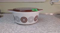 Side view of white glass casserole dish with brown and tan Bulls Eye pattern, showing double handles and lid in place.