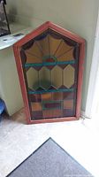 Photo showing full stained glass window leaning against wall by a desk and doorway. Window has pentagon shape and features amber, green, purple and brown colored glass with wooden reddish-brown frame.