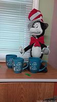3 Cat in the Hat blue coffee mugs placed in front of a 24-inch tall Cat in the Hat plastic figurine on a wood surface in front of a window.