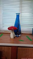 Photo of blue glass vase and red glass mushroom figurine on wooden surface with window blinds in background