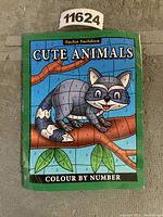 Front cover photo showing the 'Cute Animals Colour By Number' book with a raccoon illustration segmented by numbers for adding colors.
