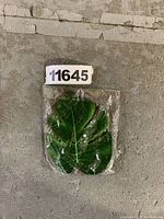 Photo of a single artificial palm leaf wrapped in clear plastic packaging laying on a worn textured surface with a lot number 11645 visible.