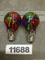 Two stained glass style LED light bulbs displayed side by side on a concrete surface with identification number in foreground.