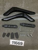 Photo displaying all physical components of the TV base stand laid out, including two V-shaped legs, two connector arms, screws, rubber pads, and assembly hex key