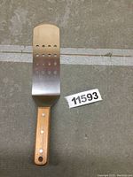 Front view of the Winco spatula turner showing perforated stainless steel blade and brown wooden handle with rivets.