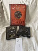 Three Game of Thrones themed books arranged on a cloth background, including 'The World of Ice & Fire,' Dothraki language book with CD, and an In Memoriam book.
