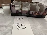 Photo showing a box containing one dozen white candles in clear glass holders with one candle missing, arranged in packaging. One empty glass candle holder placed next to the box for size comparison.