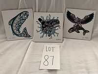 Set of three Indigenous art tiles displayed side by side on grey fabric background with lot number card in front. Tiles show animal figures in Indigenous art style.