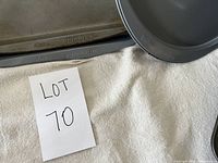 Multiple Wilton brand rectangular and round baking pans stacked on a white cloth with Lot 70 sign, showing part of the lot contents.
