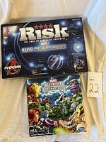 Front photo showing box covers of Marvel Risk: Marvel Cinematic Universe edition and Marvel Chess: Exiles edition games placed on white fabric.
