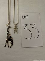 Two pendant necklaces displayed side by side on a white surface with lot number card 33. A giraffe pendant with black enamel patches and a girl pendant with crystal detail on thin silver-tone chains.