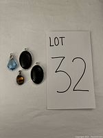 Four pendant jewelry pieces shown on white textured background including two large oval black onyx pendants, one small tiger's eye oval pendant, and one blue crystal pendant with bail.