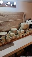 View showing multiple stacked plates, coffee mugs, egg cups, a covered pot, and creamer pitcher all in off-white ceramic with embossed floral pattern.