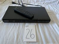 Samsung BluRay player with universal remote resting on top, front view showing Samsung and BluRay logos, power cord attached.