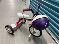 Pink and purple children's tricycle with white seat and wheels, accompanied by a purple and white helmet placed inside the tricycle's rear wheel accessory holder.