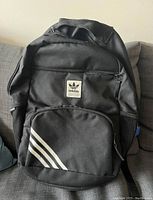 Front view of large black Adidas backpack showing front zippered pocket with three white stripes and Adidas logo patch.