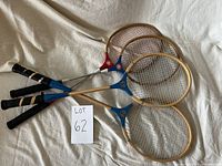 Four vintage badminton rackets displayed on a white fabric background, clearly showing wooden heads, metal shafts, and black handle grips.