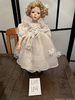 Full view of the porcelain doll standing on a wooden table wearing a white dress with puffed sleeves, floral decorations, and curly blonde hair.