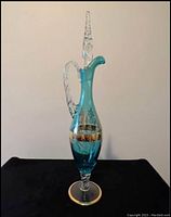 Front view of the vintage teal glass decanter decorated with gold leaf accents, showing the full height including twisted clear glass handle and stopper on top.
