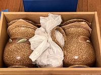 Box with two large brown textured cast iron teapots with ceramic handles wrapped in packing paper.