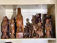 Shelf of approximately 17 Far Eastern styled wood carving figurines in various poses and finishes, showing full lot contents.