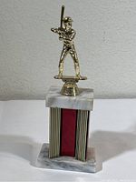 Side view of baseball trophy showing gold-tone batter figurine on a marble platform with red and gold vertical stripes on base.