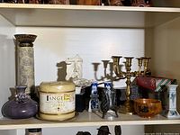 Photo showing various decorative items on a shelf including brass candelabra, brass candlestick, purple ceramic vase, and large vanilla scented candle jar.