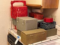 Photo showing stacked fabric covered boxes on a tiled kitchen counter, including a red box with Asian characters and handle, and several boxes with geometric and floral patterns.