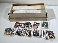 Large box filled with hundreds of unsorted baseball cards with several cards displayed in front of the box showing various players in different playing positions.