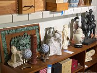The full shelf showing the variety of sculptures including Don Quixote horse rider figure, cream ceramic and stone sculptures, red carved vase, and blue-white ceramic spheres.