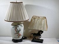 Photo of two Asian style lamps, taller white ceramic lamp with peacock design and pleated shade, smaller lamp with horse figurine base and rectangular shade