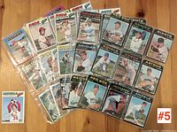 Photo of 46 baseball cards from the 1970s arranged on wood floor, showing front side mostly with some team names and pitcher/player photos visible.