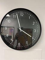 Photo of the black round IKEA wall clock hanging on a white wall showing the full clock face with hour, minute, and second hands at 4:56:55 approximately.