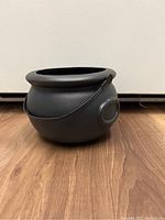 Front-angle view of a small black plastic cauldron showing its rounded shape, wide opening, and attached handle, placed on a wood floor.
