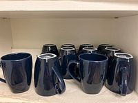 Eleven glossy cobalt blue coffee cups arranged in two rows on a shelf, some turned upside down showing the bases.