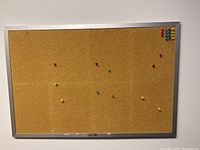 Front view of a rectangular cork bulletin board made by Stempel with a silver metal frame. Various colored push pins are pinned randomly on the board.