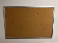 Image of a rectangular Staples bulletin board with cork surface and silver aluminum frame. Four colored push pins are pinned to the board.