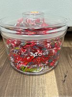 Clear round plastic holiday container filled with assorted red and silver wrapped candies. Lid is on, container has holiday text on front.