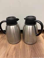 Two stainless steel coffee thermoses standing side by side on wooden floor against a light background. Each thermos has a black plastic lid with spout and black handle, showing lightly used condition.