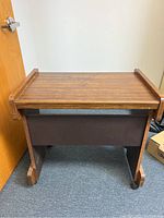 Front angled view of the medium brown wood-finish computer table with two raised side rails on top, dark brown panels, and caster wheels on legs.