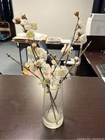 Photo shows a clear cylindrical glass vase holding several brown stems with white artificial blossoms and buds, resembling cherry blossoms, placed on a brown table surface.