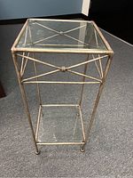 Square glass and metal accent table with two glass shelves and metal frame with X-shaped crossbar design, shown standing on carpeted flooring