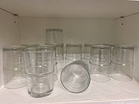 Photo showing a set of 12 clear cylindrical drinking glasses arranged in two rows, ten upright and two stacked inside others horizontally on a white shelf.