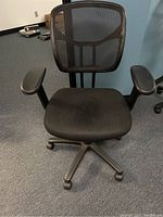 Front view of black mesh desk chair with armrests on an office floor, showing the mesh back, fabric seat, and wheeled base.